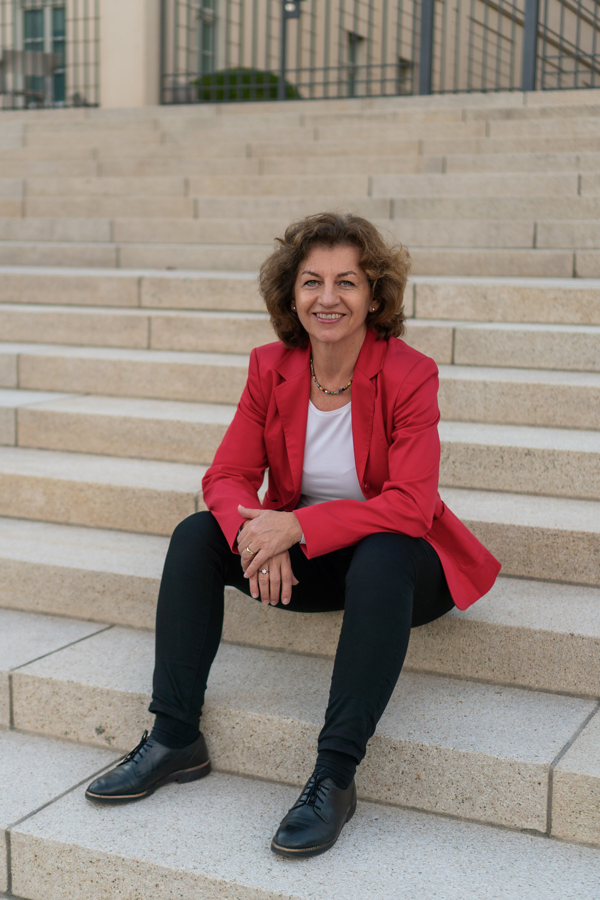 Renate Dahms im roten Jacket, schwarze Hose, auf einer hellen Treppe sitzend.